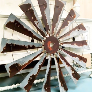 May include: A large, rustic metal windmill blade decoration with a weathered, distressed appearance. The blades are a mix of brown and gray, with visible rust and wear. The windmill has a circular center and is designed for wall mounting.