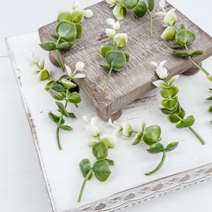 May include: A white wooden tiered stand with a decorative border. The stand is decorated with sprigs of green and white eucalyptus.