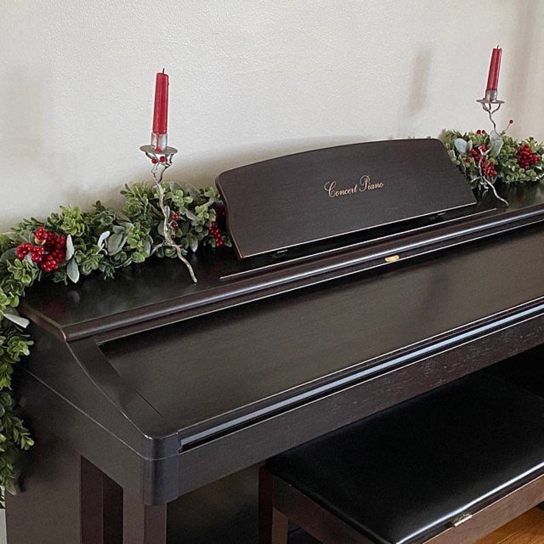 Green dark boxwood style faux greenery garland and lambs ear garland is laying on the back of a black piano being used as piano decoration. Garland is full of both pieces of greenery and has red Christmas holiday style berries distributed throughout