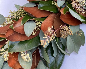 Puede incluir: Primer plano de una corona con hojas de magnolia verdes y marrones brillantes, acentuada con hojas de eucalipto verde claro y pequeños capullos beige. Ideal para la decoración del hogar.
