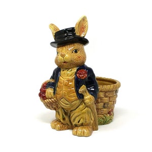 May include: A ceramic planter shaped like a rabbit wearing a black top hat and a blue jacket. The rabbit is holding a basket of red berries. The planter is a great way to add a touch of whimsy to your home decor.