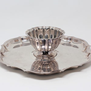 以下が含まれることがあります： 縁がギザギザの銀製のサービングトレイと、中央に置かれた小さな丸い銀製のボウル。ボウルは台座の上に置かれています。