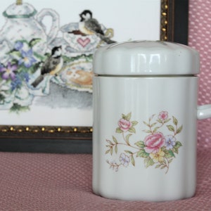以下が含まれることがあります： ピンク、黄色、青の花が描かれた白い陶器のキャニスター。キャニスターには蓋と側面にハンドルが付いています。
