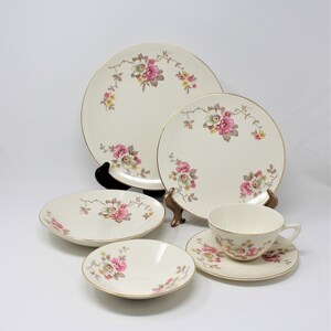 May include: A cream-coloured vintage dinnerware set with a floral design featuring pink, yellow, and green flowers. The set includes plates, bowls, a teacup, and a saucer, all with gold trim. A classic and elegant tableware collection.