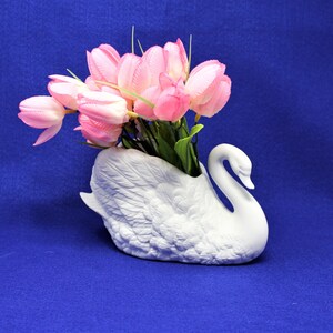 May include: A white ceramic swan vase with pink tulips. The swan vase has a detailed design and is a beautiful decorative piece.