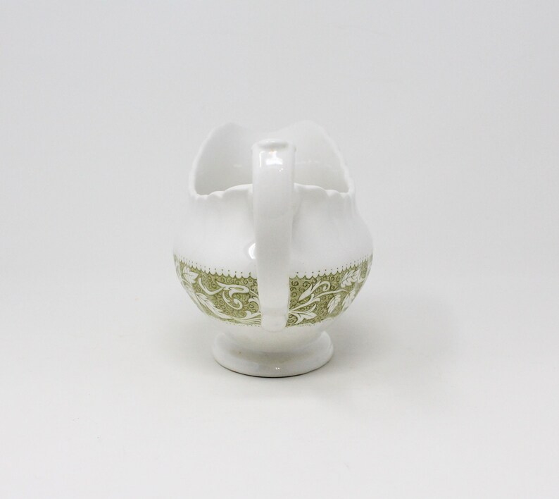 May include: White ceramic gravy boat with a green floral design. The boat has a scalloped edge and a handle.