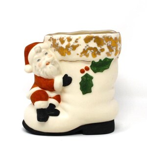 May include: A ceramic Christmas boot-shaped container with a Santa Claus figure climbing on the side. The boot is off-white with gold accents, holly, and red berries. The Santa figure is wearing a red suit and hat.