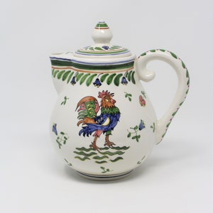May include: A white ceramic teapot with a rooster design. The rooster is painted in shades of blue, green, orange, and red. The teapot has a green and white lid and a green and white handle.
