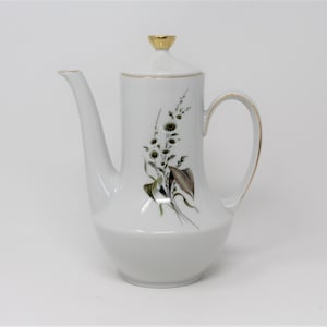 Puede incluir: Cafetera de cerámica blanca con ribete dorado y diseño floral. Las flores son blancas con hojas verdes.
