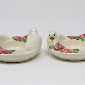 May include: Two white ceramic candle holders shaped like doves with pink roses and green leaves. The doves are facing each other and have yellow beaks.