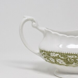 May include: White ceramic gravy boat with a green floral design. The boat has a curved handle and a scalloped edge.