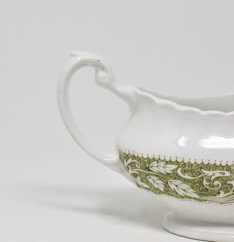 May include: White ceramic gravy boat with a green floral design. The boat has a curved handle and a scalloped edge.
