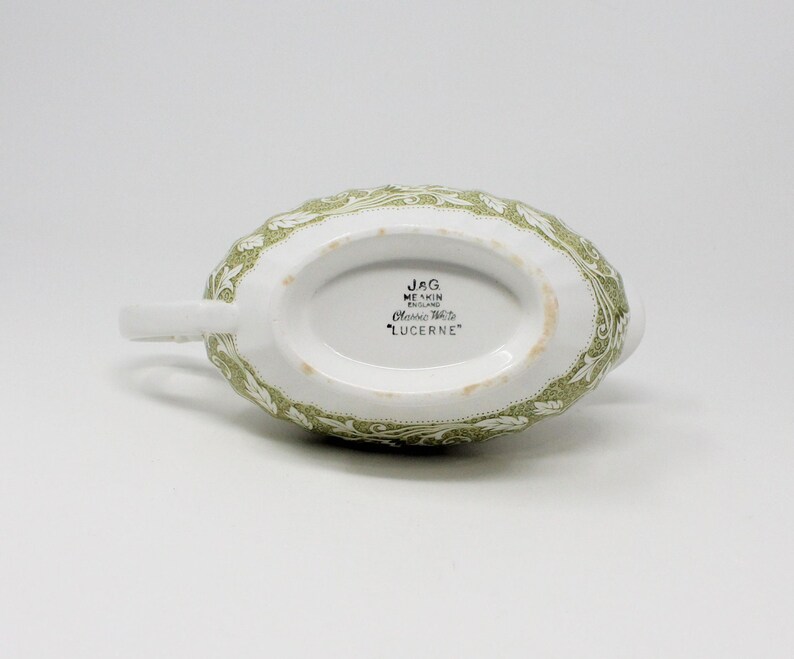 May include: A white ceramic gravy boat with a green floral pattern. The bottom of the boat has the text "JAG MEAKIN ENGLAND Classic Ware 'LUCERNE'" printed on it.