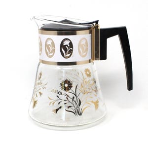 May include: A vintage glass coffee carafe with a black handle and lid. The carafe features a gold and white band with floral designs and gold floral patterns on the glass. The carafe is a classic retro kitchenware item.