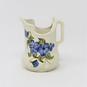 May include: A white ceramic creamer with a floral design. The creamer has a unique shape with a wide opening and a handle on the side. The flowers are blue and green and are painted on the front of the creamer.