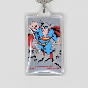 May include: A clear plastic keychain featuring a comic book illustration of Superman flying with a red cape and blue suit. The text "© DC COMICS INC. 1983" and "BACK-UP TROY, MI" is printed on the keychain.