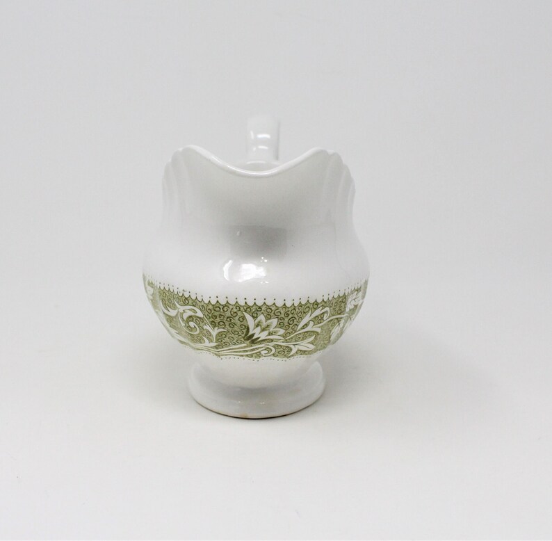 May include: White ceramic creamer with a green floral design around the base. The creamer has a handle and a rounded shape.