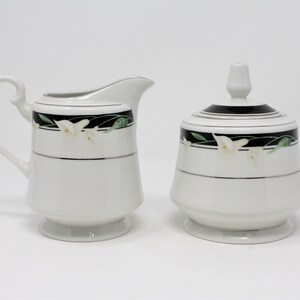 Puede incluir: Un juego de crema y azúcar blanco con diseños florales negros y verdes. La jarra tiene un asa y un pico. El azucarero tiene una tapa.