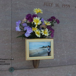 Puede incluir: Una foto dorada de una pareja de pie en una playa se muestra en un jarrón dorado con un ramo de flores moradas, amarillas y blancas. El texto "JULY 16, 1959 + JULY" es visible en la pared detrás del jarrón.