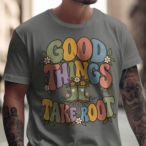 May include: A grey t-shirt featuring the text "GOOD THINGS TAKE ROOT" in large, colorful, bubble-style letters. The design includes a central illustration of green rain boots filled with flowers, surrounded by small white daisies and green leaves.