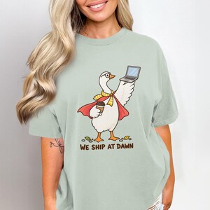 Può includere: Una t-shirt verde salvia con la grafica di un'oca bianca che indossa un mantello rosso, una sciarpa gialla e degli occhiali. L'oca tiene in mano un portatile e una tazza di caffè. Sotto l'illustrazione, il testo WE SHIP AT DAWN in marrone.