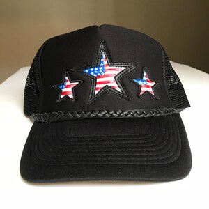 May include: Black trucker hat with a black braided band and three stars. The stars are red, white, and blue and feature the American flag design.