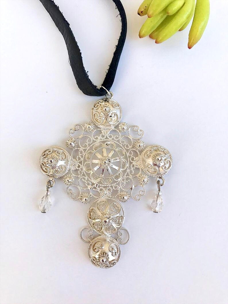 May include: A silver filigree pendant necklace with a black ribbon. The pendant features a large, intricate design with four round, ornate elements and two crystal drops.
