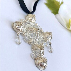 May include: A silver filigree pendant with a cross shape and crystal accents hanging from a black ribbon.