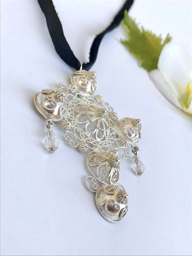 May include: A silver filigree pendant with a cross shape and crystal accents hanging from a black ribbon.