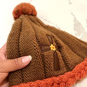 May include: Brown knitted beanie with a brown cross and a yellow flower embroidered on the front. The beanie has a brown pom pom on top and an orange knitted brim.