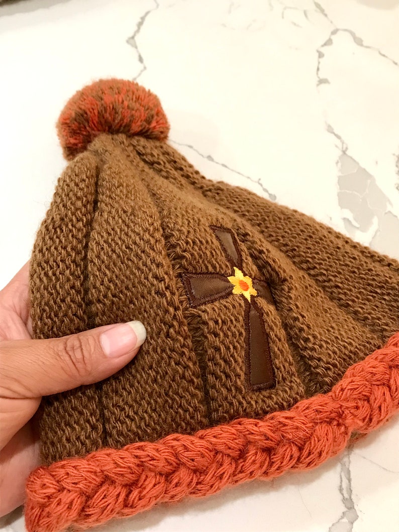 May include: Brown knitted beanie with a brown cross and a yellow flower embroidered on the front. The beanie has a brown pom pom on top and an orange knitted brim.