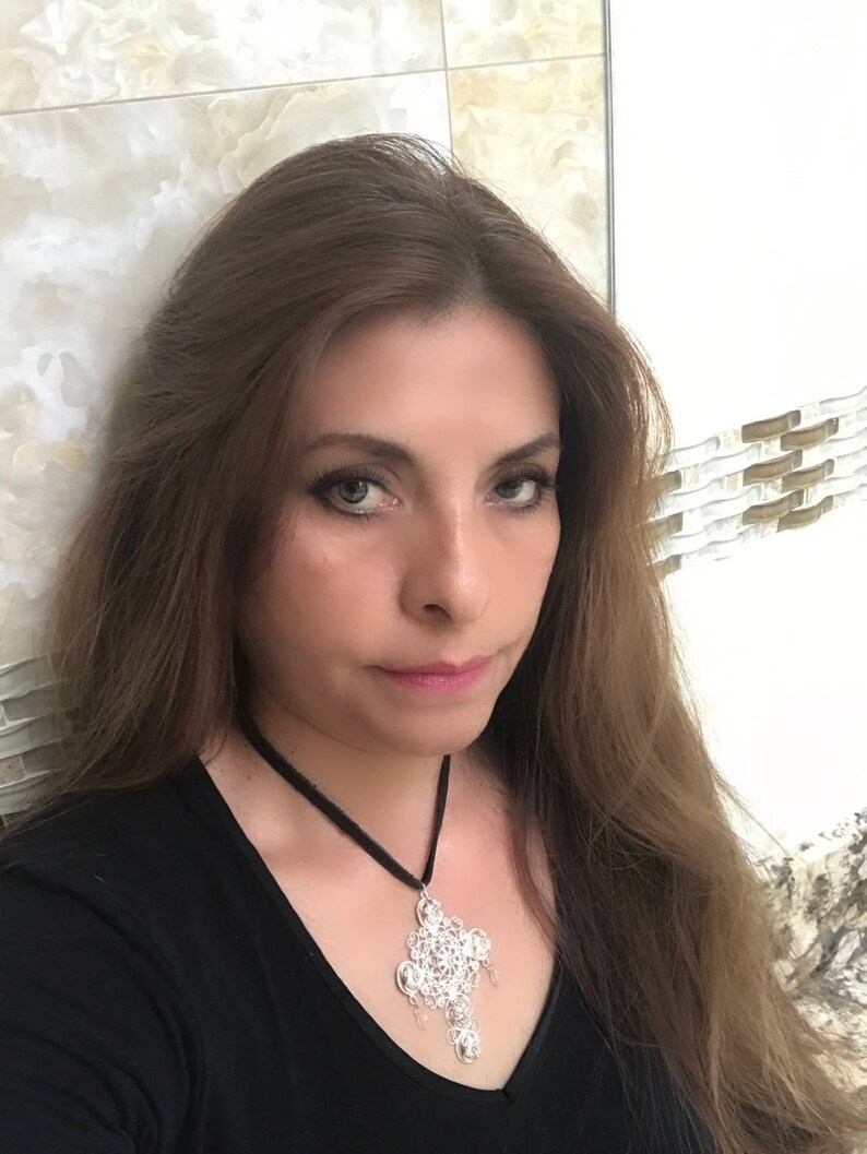 May include: A woman with long brown hair is wearing a black shirt and a silver necklace with a large pendant. The pendant is a cross-shaped design with intricate details.