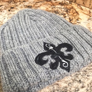 May include: Grey knitted beanie with a black embroidered fleur-de-lis design.