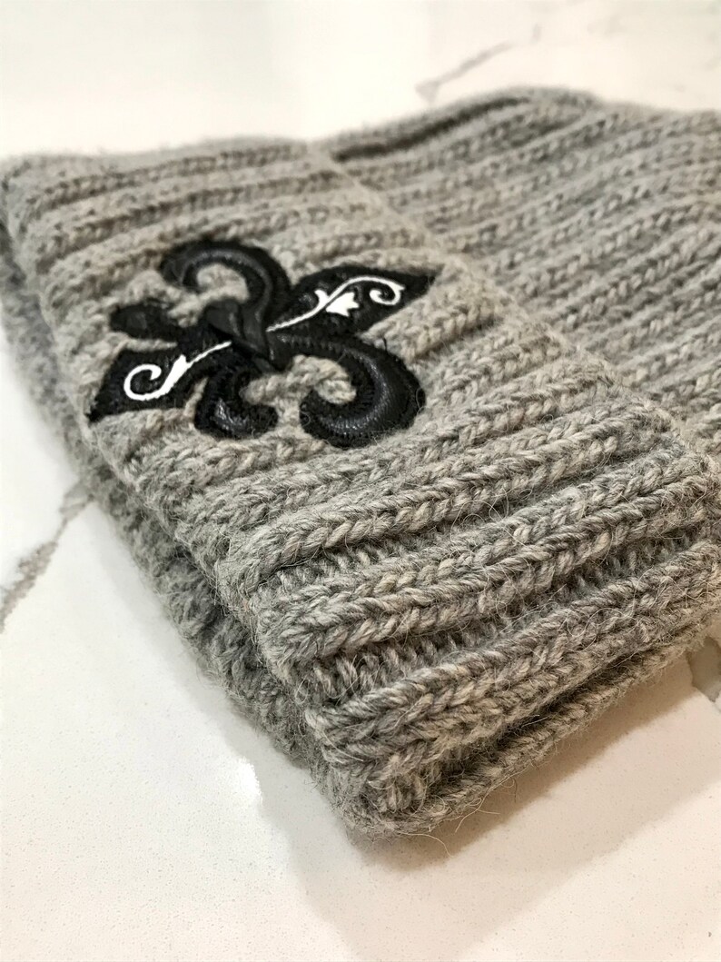 May include: Grey knitted beanie with a black embroidered fleur-de-lis design.
