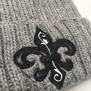 May include: Grey knitted beanie with a black and white fleur-de-lis embroidered on the front.