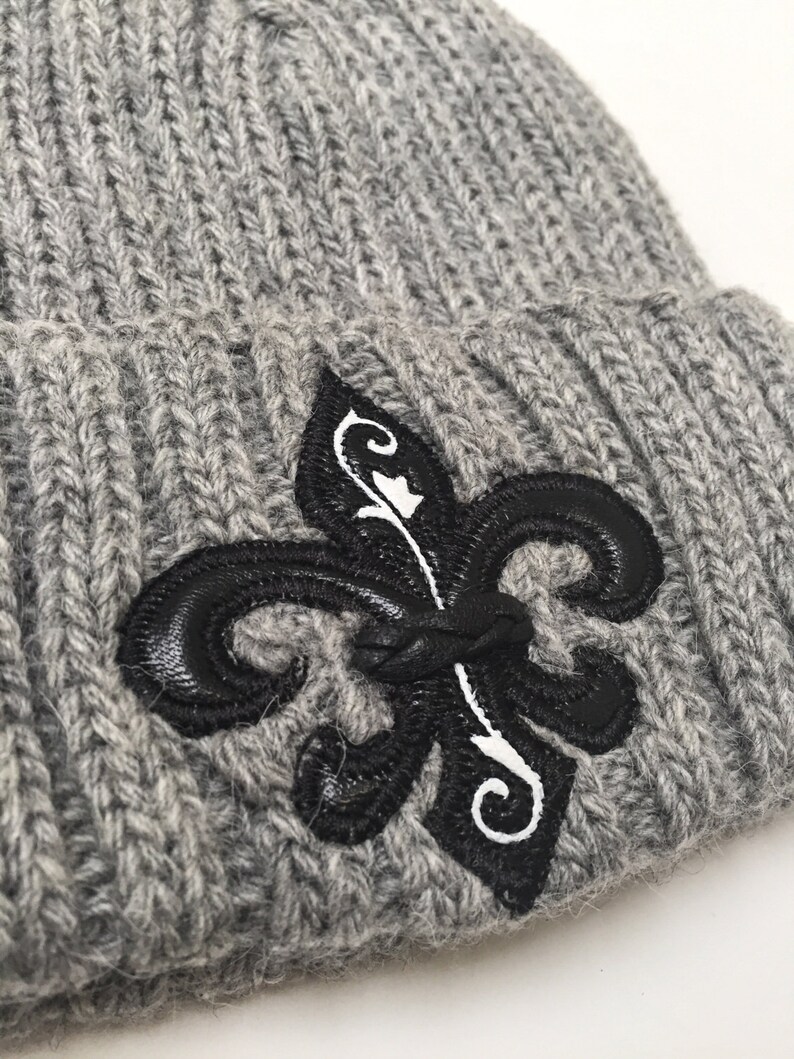 May include: Grey knitted beanie with a black and white fleur-de-lis embroidered on the front.