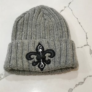 May include: Grey knitted beanie with a black and white fleur-de-lis embroidered design.