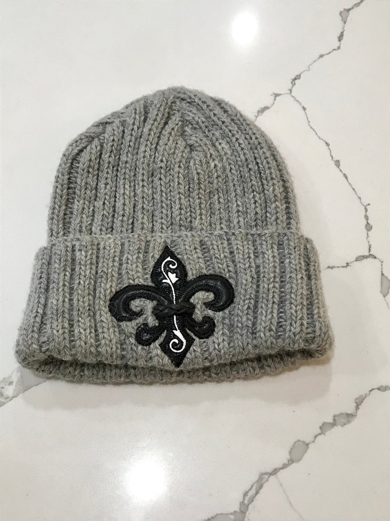 May include: Grey knitted beanie with a black and white fleur-de-lis embroidered design.