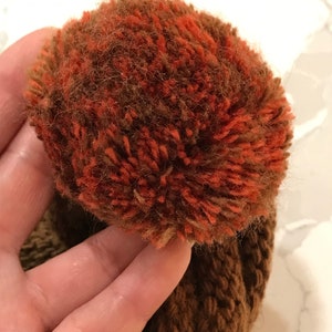May include: A brown knit hat with a large, fluffy pompom made of brown and orange yarn.