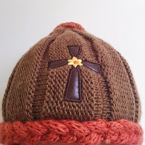 May include: Brown knitted beanie with a brown leather cross and a yellow flower embroidered on the front. The beanie has a red knitted brim.