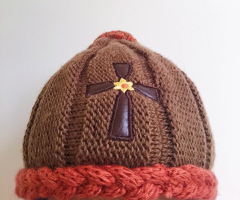 May include: Brown knitted beanie with a brown leather cross and a yellow flower embroidered on the front. The beanie has a red knitted brim.