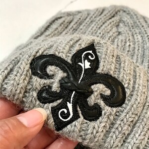 May include: Grey knitted beanie with a black leather fleur-de-lis patch.