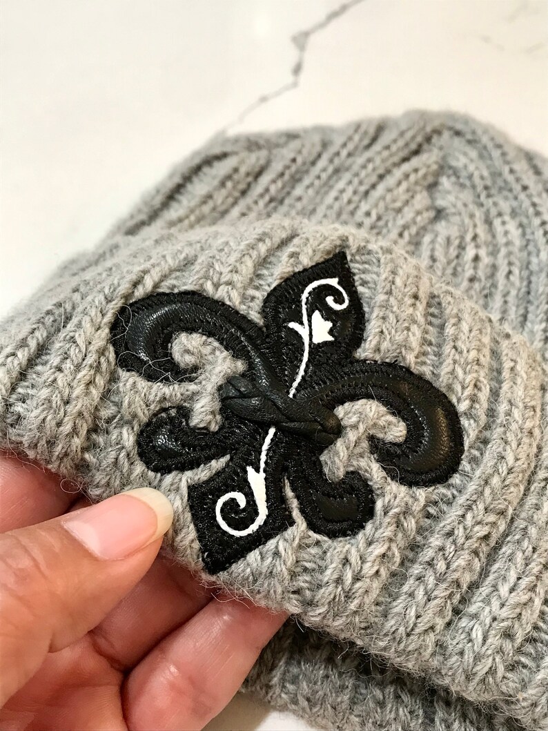 May include: Grey knitted beanie with a black leather fleur-de-lis patch.