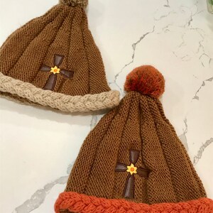 May include: Two knitted winter hats with pom poms. One hat is brown with a tan brim and a brown cross with a yellow flower. The other hat is brown with an orange brim and a brown cross with a yellow flower.