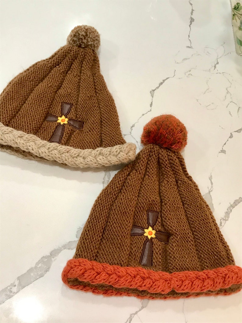 May include: Two knitted winter hats with pom poms. One hat is brown with a tan brim and a brown cross with a yellow flower. The other hat is brown with an orange brim and a brown cross with a yellow flower.