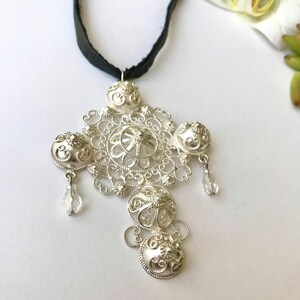 May include: A silver filigree pendant necklace with a black ribbon. The pendant is a cross shape with intricate details and clear crystal accents.