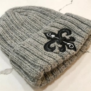 May include: Grey knitted beanie with a black embroidered fleur-de-lis design.