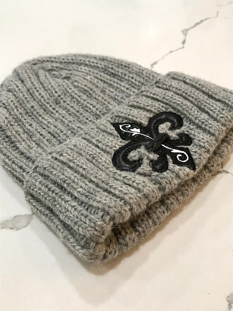 May include: Grey knitted beanie with a black embroidered fleur-de-lis design.