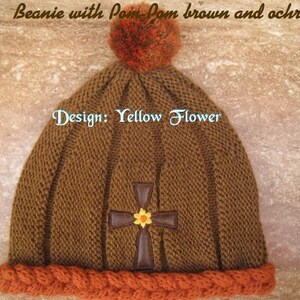 May include: Brown knit beanie with a pom pom and a cross design with a yellow flower. The beanie has an orange braided brim. The text "Design: Yellow Flower" is visible on the beanie.