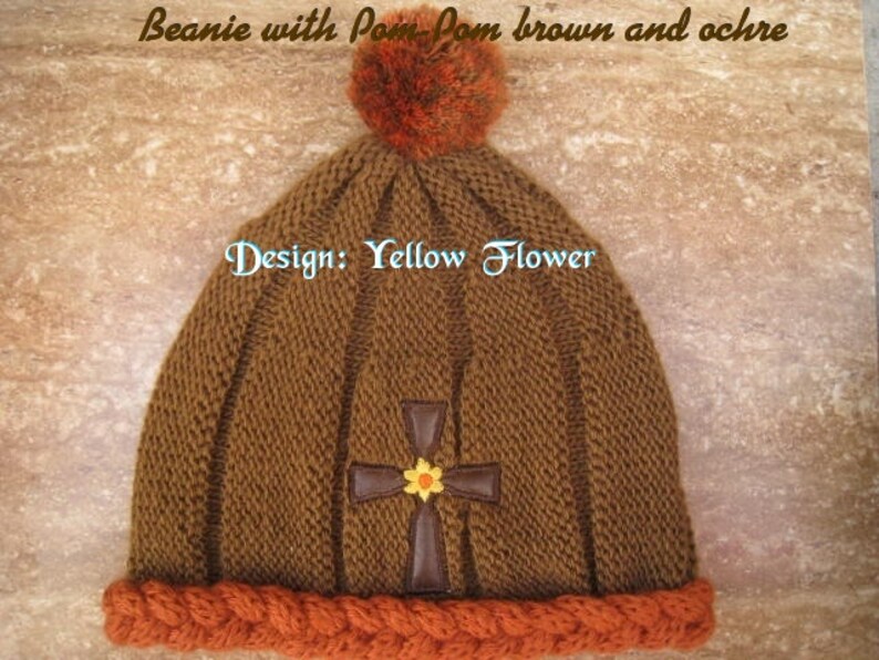 May include: Brown knit beanie with a pom pom and a cross design with a yellow flower. The beanie has an orange braided brim. The text "Design: Yellow Flower" is visible on the beanie.